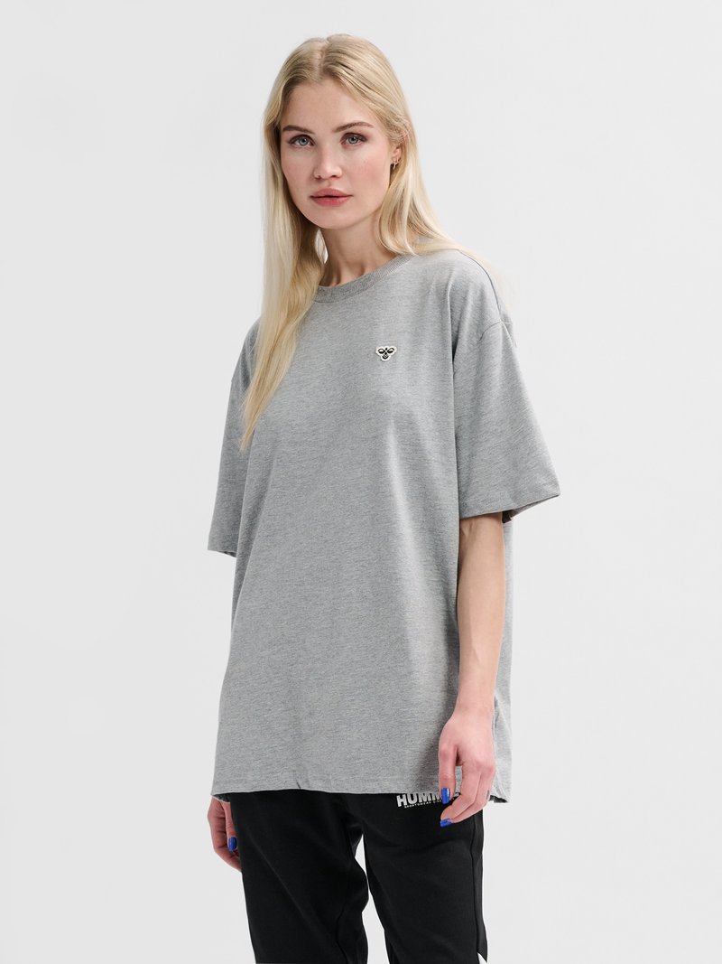 Femme blonde portant un t-shirt gris ample avec un petit logo d’abeille et un pantalon noir, debout devant un fond clair unicolore.
