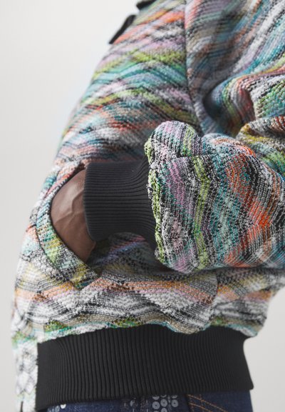Veste en tricot avec un motif zigzag multicolore dans des tons de gris, vert, orange et rose. Poignets noir côtelé et détail de poche intégré.