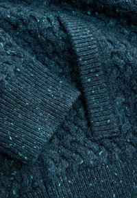 Tirkīza adīta džemperi audums ar strukturētu ribi un kabeļu rakstiem, ar gaišām specklēm, lai piešķirtu papildu vizuālo detaļu.