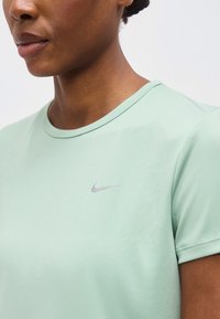 Κοντινό πλάνο ενός ατόμου που φοράει ένα ανοιχτό πράσινο μπλουζάκι Nike με στρογγυλή λαιμόκοψη και μικρό γκρι λογότυπο Nike στο στήθος.