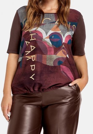 Kvinde iført en vinrød og brun top med abstrakt fugledesign og teksten "Happy", kombineret med mørkebrune læderbukser.