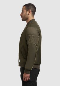 Olivgrüne Bomberjacke mit einer Reißverschlusstasche am Ärmel, gerippten Bündchen und Saum sowie einer glatten, glänzenden Oberfläche.