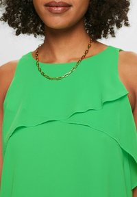 Top senza maniche verde con strati arricciati, realizzato in tessuto leggero. Accentuato da una collana con catena dorata. Vista ravvicinata.