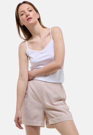 Femme à la peau claire et aux cheveux raides portant un caraco blanc et un short beige rayé, posant devant un fond uni.