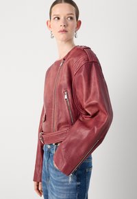 Raudona odinė striukė su lygiu paviršiumi, priekiniais ir šoniniais užtrauktukais, ir prigludusia forma. Dėvima su mėlynomis džinsomis.