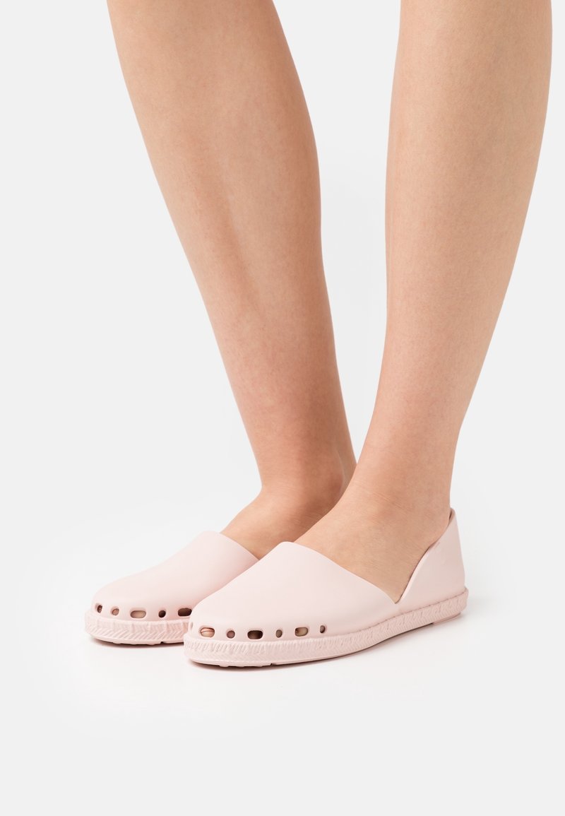 Paire de jambes portant des chaussures légères roses avec des trous de ventilation le long des semelles, debout sur un fond blanc.