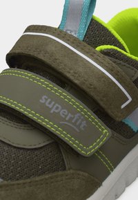 Κοντινό πλάνο παιδικού αθλητικού παπουτσιού σε ελιά πράσινο, με δύο αυτοκόλλητα λουράκια βέλκρο, ραφές σε νέον πράσινο και φωτεινή κίτρινη επένδυση στο εσωτερικό.