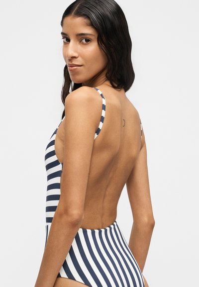 Femme aux longs cheveux foncés portant un maillot de bain dos nu rayé bleu marine et blanc, regardant par-dessus son épaule sur un fond uni.