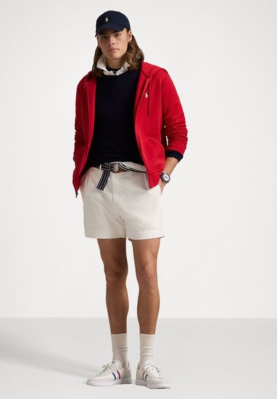 Camisola com fecho éclair vermelha, suéter azul-marinho, calções brancos e sapatilhas brancas com detalhes às riscas. Cinto claro e boné azul-marinho completam o look.