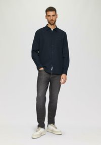 Camicia button-up blu scuro con taschino, abbinata a jeans grigi e sneakers bianche. La camicia presenta un colletto classico e maniche lunghe.