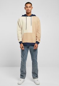 Hoodie v krémovo-hnedých farbách s námorníckymi akcentami. Obsahuje predné vrecko, sťahovacie šnúrky a mäkký materiál. Nosené s svetlými džínsami.