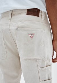 Pantalones cargo beige con dos bolsillos traseros, un parche con logo en forma de triángulo y una etiqueta de cuero marrón texturizado en la cintura. Cuenta con detalles de herrajes metálicos.