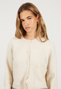 Crème kleurige cardigan van zacht materiaal, met een ronde halslijn, vijf knopen en twee voorkeurszakken.