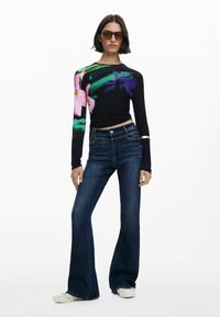 Juoda ilgomis rankovėmis palaidinė su spalvingu gėlėtu raštu ant vienos rankovės, derinama su tamsiai mėlynomis platėjančiomis džinsomis ir baltomis šlepetėmis.