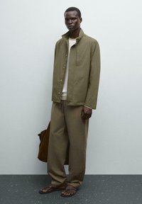 Giacca leggera verde oliva con colletto alto, tasca sul petto, abbinata a pantaloni beige e sandali marroni. Design minimalista, vestibilità rilassata.