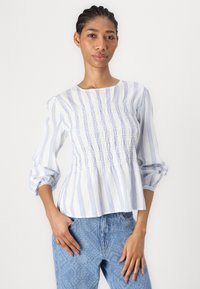 Hvit bluse med blå vertikale striper, med rynket tekstur langs kroppen og trekvartlange ermer. Kombinert med lyseblå mønstrede bukser.