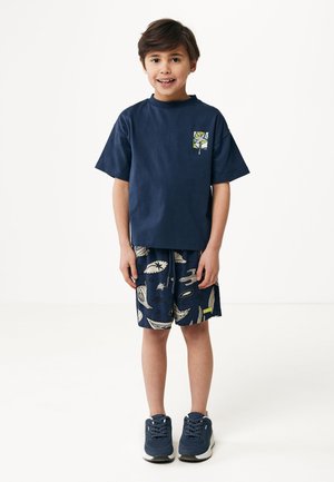 T-shirt en coton bleu marine à manches courtes avec une poche frontale ; associé à un short bleu marine à motifs avec des accents blancs et des baskets bleues.