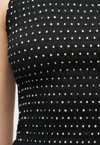 Μαύρο αμάνικο τοπ με οριζόντιες σειρές μικρών λευκών πουα, φορεμένο από άτομο με ορατό αριστερό βραχίονα.