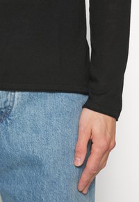 Rechte Hand ruht neben hellblauen Jeans, getragen wird ein schwarzes langärmliges Oberteil mit einem eng anliegenden Bündchen.