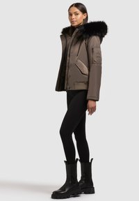 Braune isolierte Jacke mit einer Kapuze aus schwarzem Kunstfell, ausgestattet mit einem Reißverschluss und Druckknopfverschluss, kombiniert mit schwarzen Leggings und auffälligen schwarzen Stiefeln.