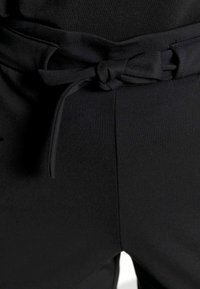 Zwarte broek van een gladde stof, met een strikdetail op de tailleband en een vlakke voorkant. Geen zichtbare zakken.
