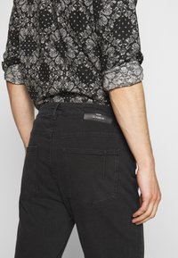 Černé džíny z džínoviny s klasickým střihem, s koženou etiketou na pasu a dvěma zadními kapsami, vystavené vedle vzorované košile.