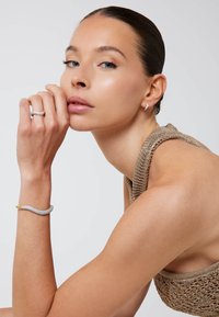 Metallic ring with a wave design, silver bracelet with gold accents, and a textured beige knit top. Smooth skin and natural makeup.