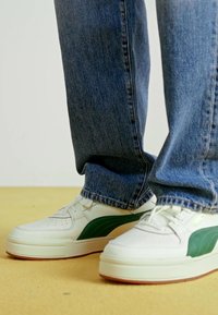 Személy fehér sneakersben, zöld csíkkal, kék farmernadrágban áll egy sárga padlón.