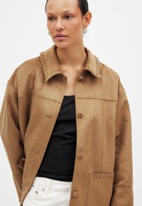 Pruunist seemisjakist, millel on kraega kaelus, nööbidega sulgemine ja kootud detail krae äärtes. Taskutel on sobiv õmblus.