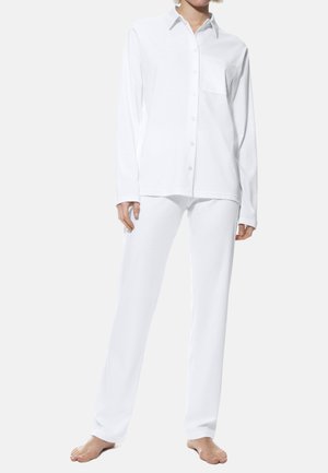 Persoon die op blote voeten staat en een geheel wit outfit draagt met een blouse met lange mouwen en knopen en bijpassende rechte broek.