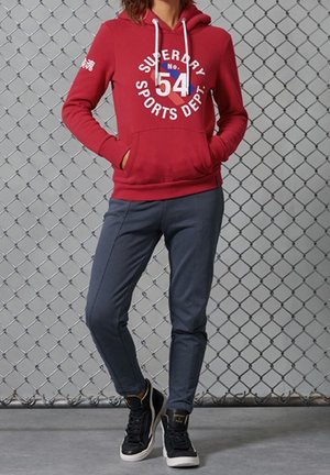 Felpa rossa con grafica bianca "SUPERDRY 54 SPORTS DEPT" sul davanti, abbinata a pantaloni jogger grigi aderenti e sneakers nere alte.