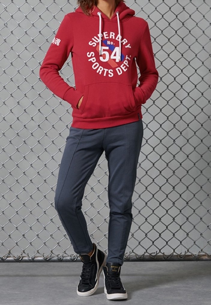 Sudadera roja con gráfico blanco "SUPERDRY 54 SPORTS DEPT" en la parte delantera, combinada con joggers gris ajustados y zapatillas altas negras.