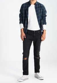 Neformální outfit zahrnující modrou károvanou košili, bílou pruhovanou košili s dlouhým rukávem, černé roztrhané džíny a bílé tenisky.