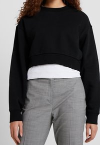 Sudadera negra recortada con mangas largas, cuello redondo y textura suave, superpuesta a una camiseta blanca, combinada con pantalones de cuadros grises.