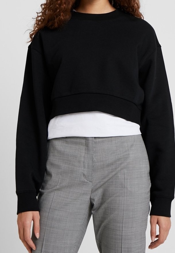 Sudadera negra recortada con mangas largas, cuello redondo y textura suave, superpuesta a una camiseta blanca, combinada con pantalones de cuadros grises.