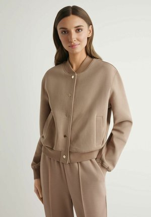 Kobieta w beżowej, zapinanej na guziki bomberce z prążkowanymi mankietami i dołem, w komplecie z dopasowanymi, wysokim stanem spodniami, stojąca na tle jednolitego tła.