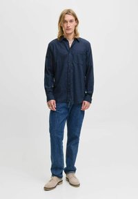 Man with long blond hair wearing navy button-up shirt, blue jeans, and beige shoes standing against plain white background.