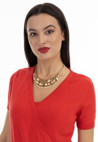 Arany nyaklánc rétegezett levélformájú díszítésekkel, amelyet egy testhez álló, piros, kötött anyagból készült, rövid ujjú, V nyakú felsővel viselnek.