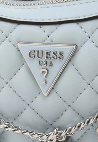 Lichtblauw, gequilt synthetisch leer met een driehoekig zilveren embleem met de tekst "GUESS USA" en een vraagteken symbool. Kettingdetail zichtbaar.