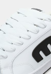 Λευκό αθλητικό παπούτσι με δερμάτινο επάνω μέρος με υφή, διαθέτει επίπεδα λευκά κορδόνια και μαύρη λογότυπο στην πλευρά. Παρουσιάζεται λεπτομέρεια ραφής.