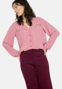 Różowa bluzka z długim rękawem z dekoltem w serek, z koronkowymi detalami na klatce piersiowej i zapięciem na guziki, zestawiona z burgundowymi spodniami.