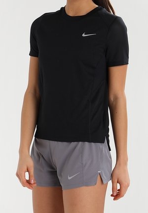 Kobieta ubrana w czarną krótki rękaw koszulkę Nike i szare sportowe szorty Nike, stoi z ramionami luźno opuszczonymi na tle jednolitego tła.