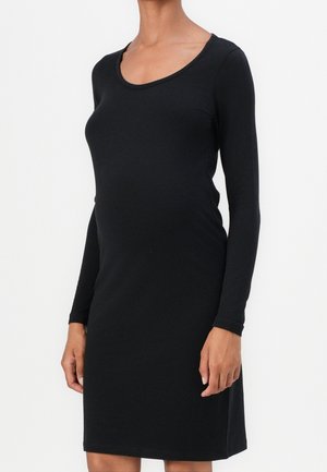 Femme portant une robe noire moulante à manches longues avec un col rond, debout devant un fond blanc uni.
