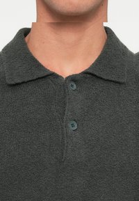 Polo shirt grigio scuro realizzato in tessuto testurizzato. Presenta un colletto e una piastra con tre bottoni di colore scuro. Design aderente.