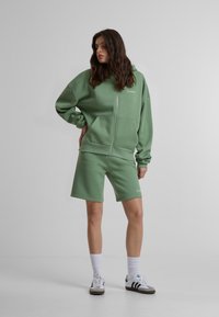 Veste à zip vert clair avec poches kangourou, associée à un short assorti. Fabriquée en tissu doux, elle présente un logo discret et des poignets côtelés.