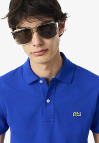 Blå bomullspolo med strukturerad yta, treknapps placket och en liten grön broderad krokodil-logga på vänster bröst.