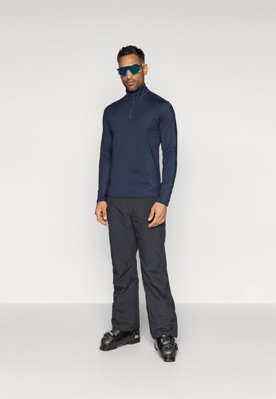 Camisa de manga comprida azul-marinho com fecho, combinada com calças de esqui pretas e botas de esqui pretas. A camisa tem um colarinho alto e um design ajustado.