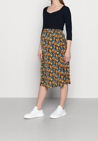 Těhotenské šaty s černým topem a květovanou sukní v oranžové, modré a zelené barvě, s délkou do kolen. Vyneseno s bílými teniskami.