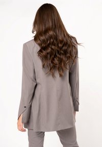 Gray tailored blazer with long sleeves, single-button cuffs, and a slightly oversized fit, paired with matching trousers. Smooth fabric texture.