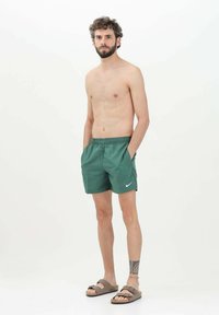 Pantaloni da bagno verdi con cintura elastica e tasche laterali, con un piccolo logo Nike bianco. Indossati con sandali marroni.